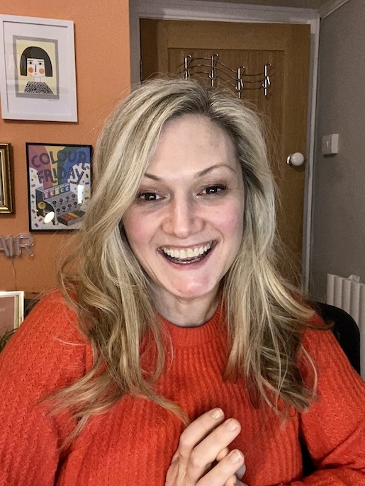 Photo of a woman with long blond hair smiling, wearing an orange jumper.